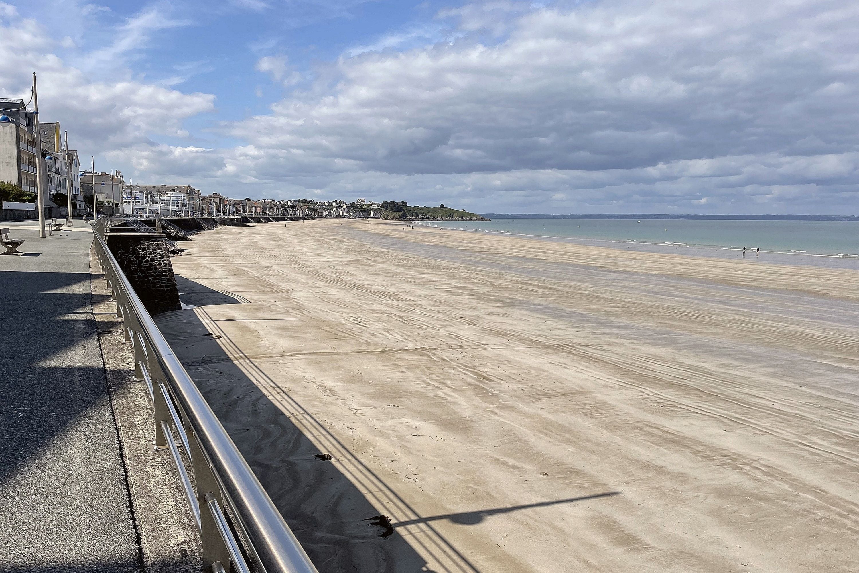 Um calçadão costeiro com uma praia de areia, áreas de estar e uma vista distante de edifícios e colinas.