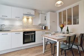 A modern kitchen with white cabinetry, a built-in oven, and a dining area with a table set for two. - Roz-Val - Appartement avec jardin proche de la mer (Pléneuf-Val-André)