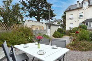 An outdoor patio with a table set for two, surrounded by greenery and a building in the background. - Roz-Val - Appartement avec jardin proche de la mer (Pléneuf-Val-André)