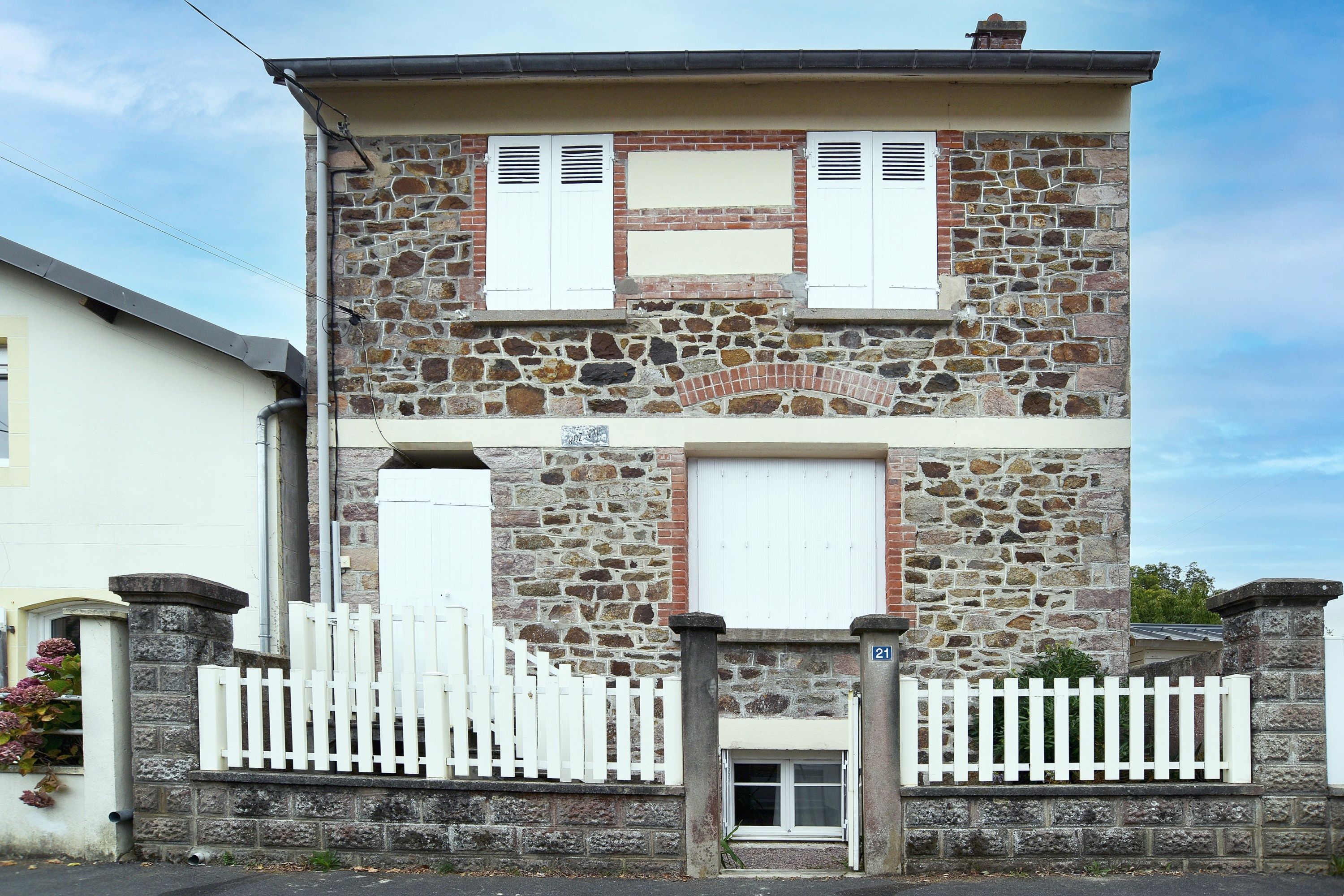 A two-story stone building with a white picket fence, a small entrance door, and a number plaque.
