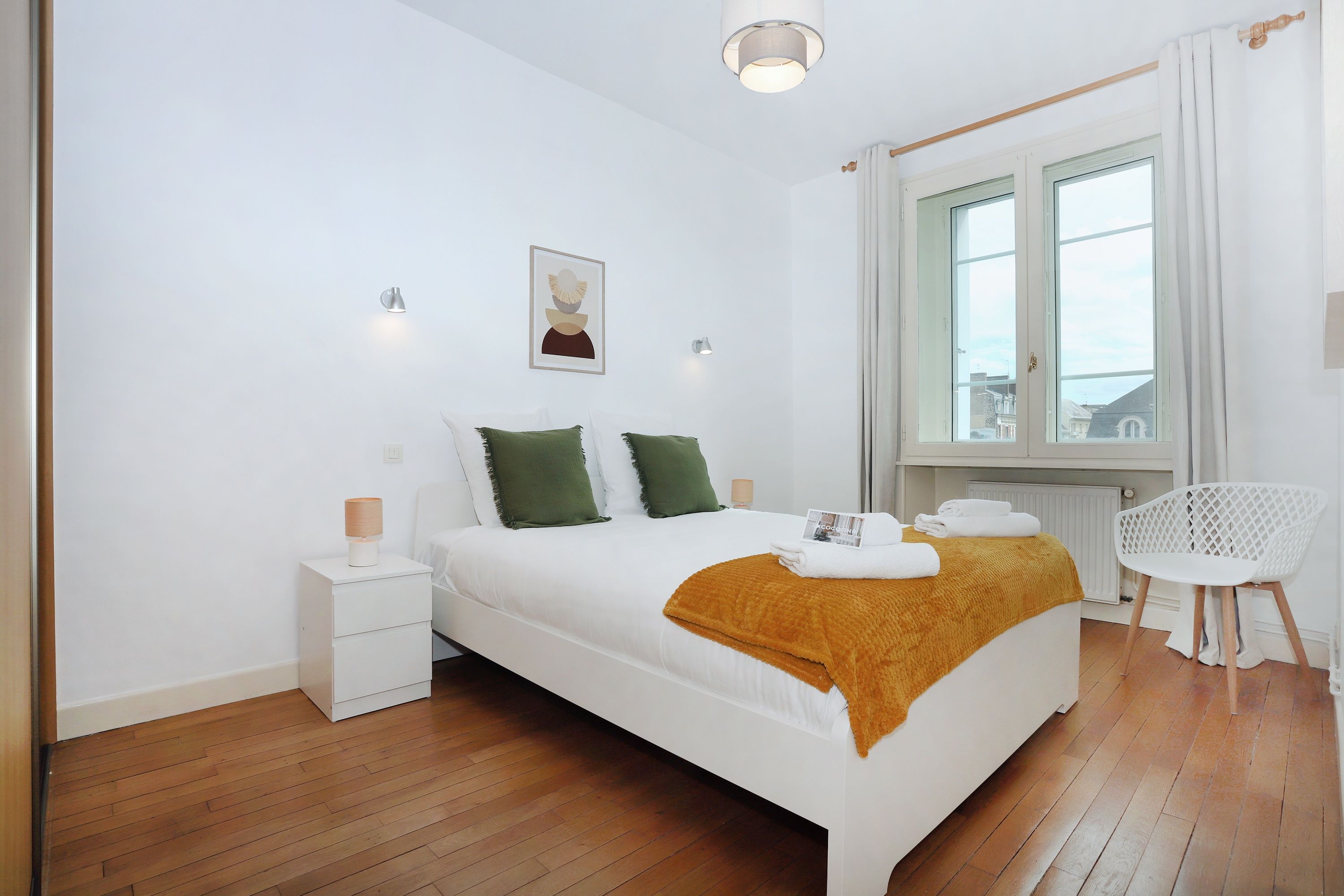 Ein Hotelzimmer mit Bett, Nachttisch, Stuhl, einem Fenster mit Vorhängen und Blick nach draußen.