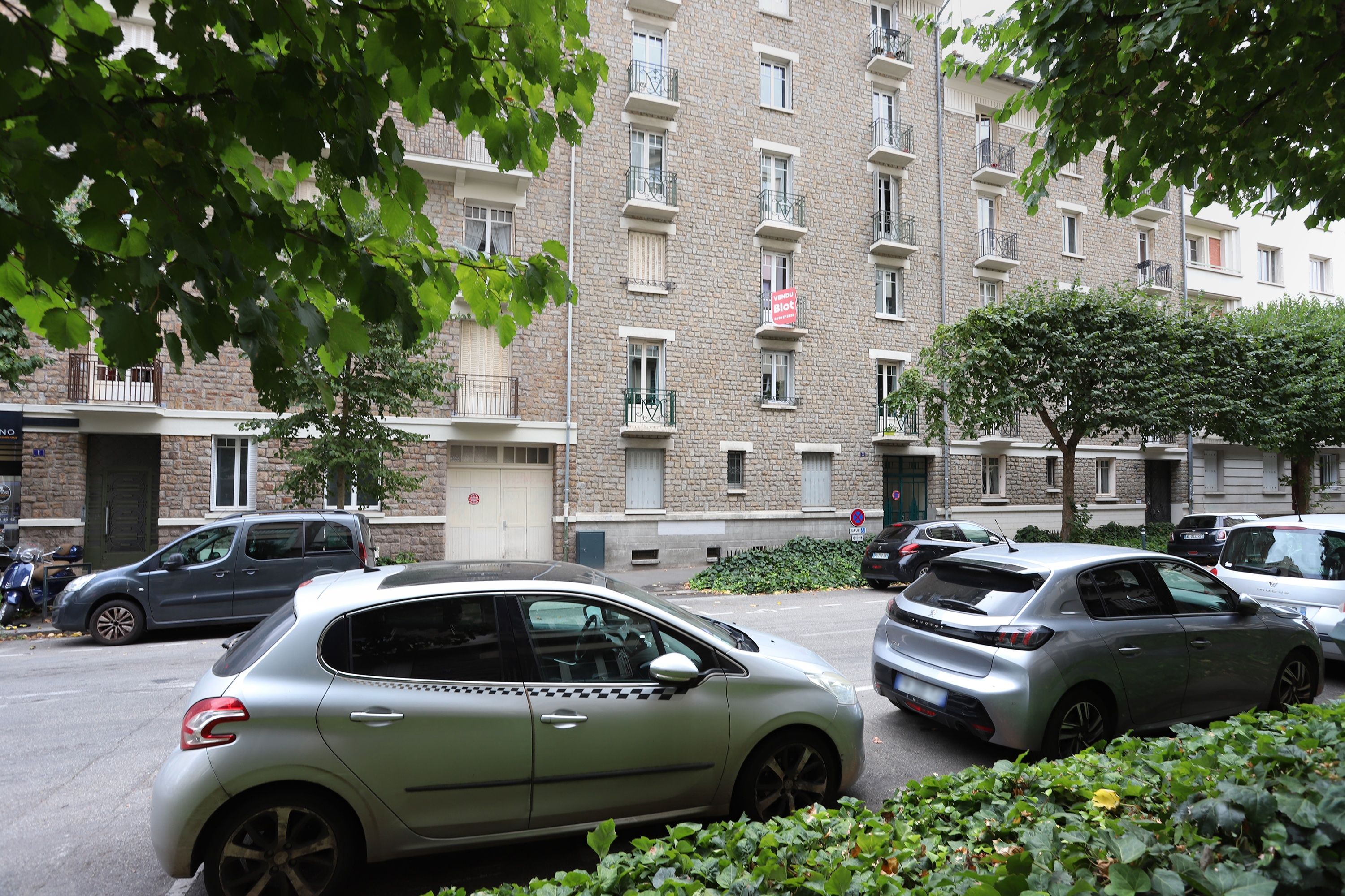 A multi-story residential building with a stone facade, parked cars, and greenery.