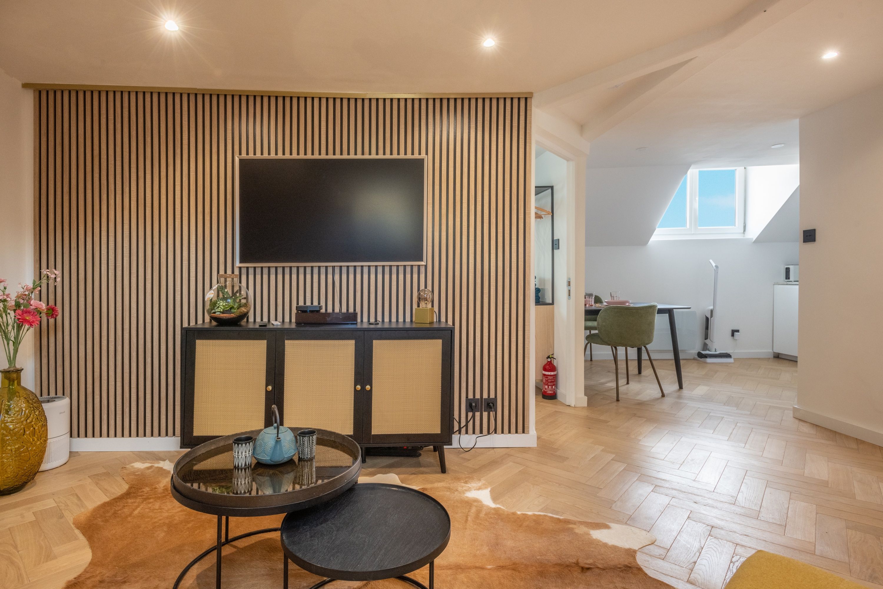 A modern living room with a flat-screen TV mounted on a wooden panel wall, a round dining table with chairs, and a kitchen area visible through an open doorway.