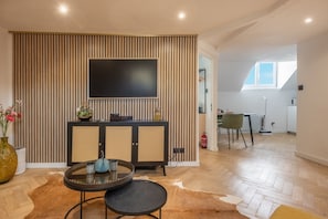 Ein modernes Wohnzimmer mit einem Flachbildfernseher an einer Wand aus Holzpaneelen, einem runden Esstisch mit Stühlen und einer durch eine offene Tür sichtbaren Küchenzeile.