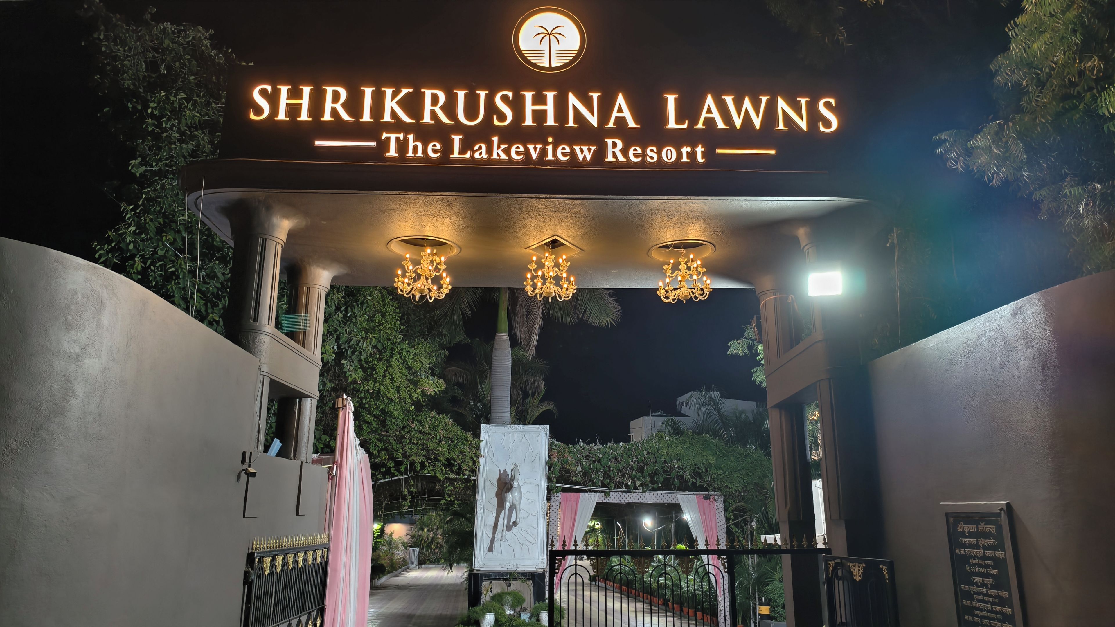La entrada a Shrikrushna Lawns The Lakeview Resort, con un letrero sobre la puerta, candelabros y un camino que conduce al interior de la propiedad.