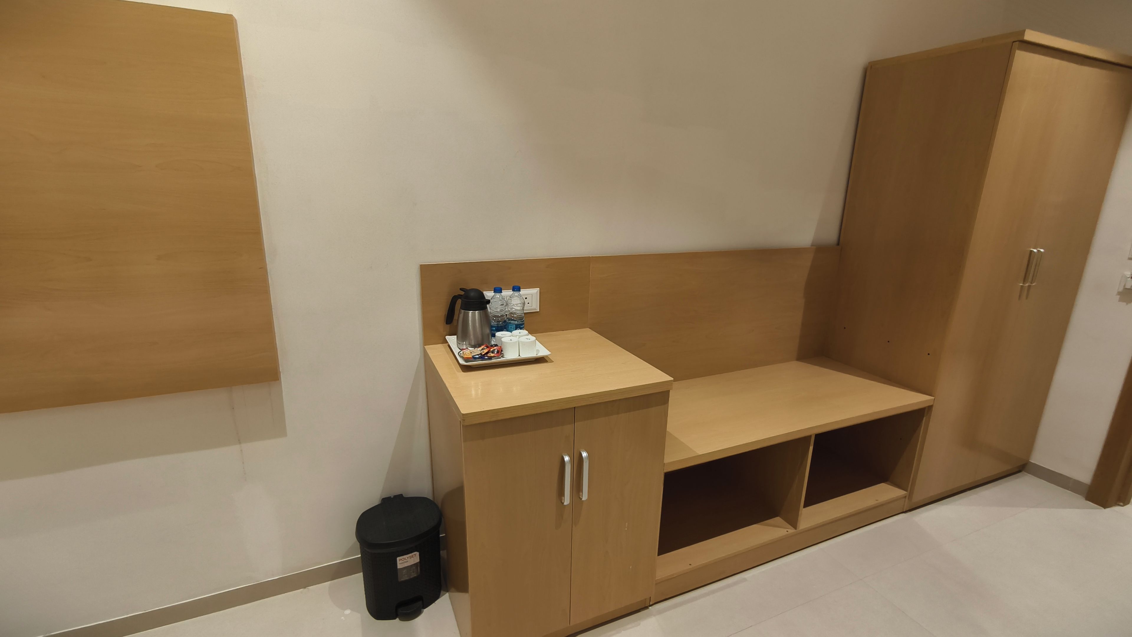 Un cuarto pequeño y cerrado con un banco de madera, un bote de basura y una mesa con una botella de agua, un termo y un plato de aperitivos.