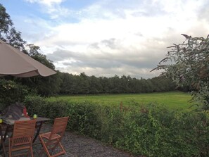 Área externa com mesas e cadeiras, um guarda-sol e vista para um campo gramado e árvores.