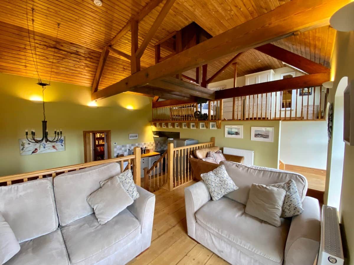 A cozy living room with two sofas, a wooden ceiling, and a kitchen area in the background.