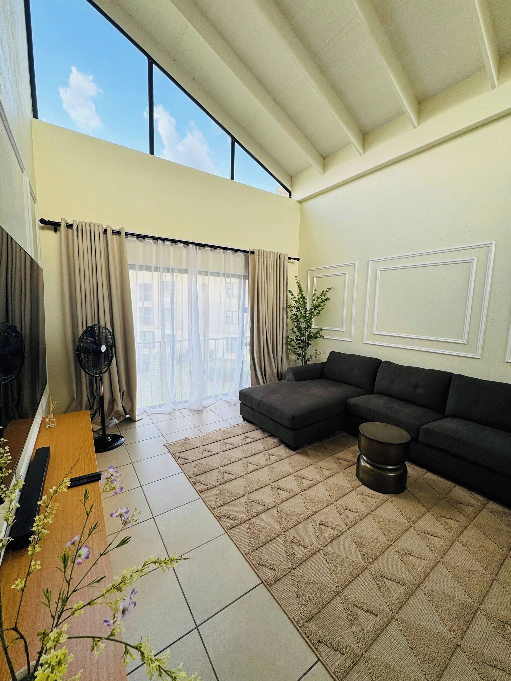 A living room with a sectional sofa, a fan, a potted plant, and a view of the sky through large windows.