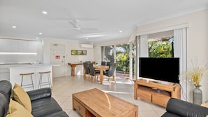 A modern living room with a sofa, coffee table, and TV. There is a dining area with a table and chairs. The room has large windows and a balcony. - Sanctuary Lake Apartment 67 - Beachside Oasis in Currumbin - Min. 3 Night Stays - Linen Provided (Currumbin)