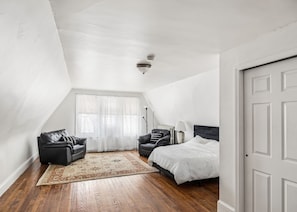 A bedroom with a sloped ceiling, a bed, two chairs, a floor lamp, and a large window with sheer curtains.