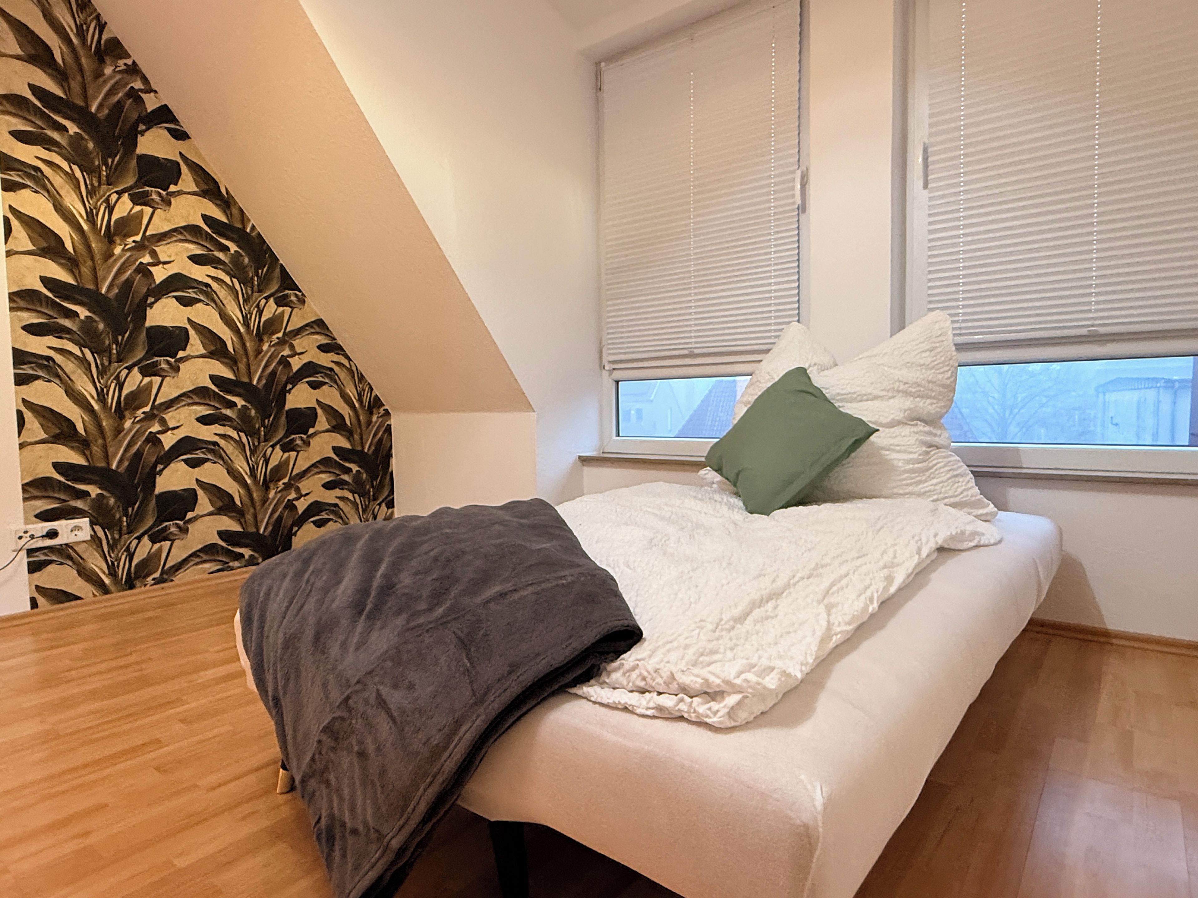 Un dormitorio con cama, ventana con persianas, almohada verde y pared con diseño de hojas.