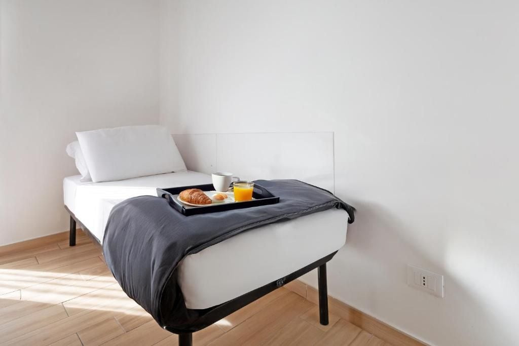 Una cama individual con colchón blanco, cabecero negro y una bandeja con croissants y jugo de naranja.