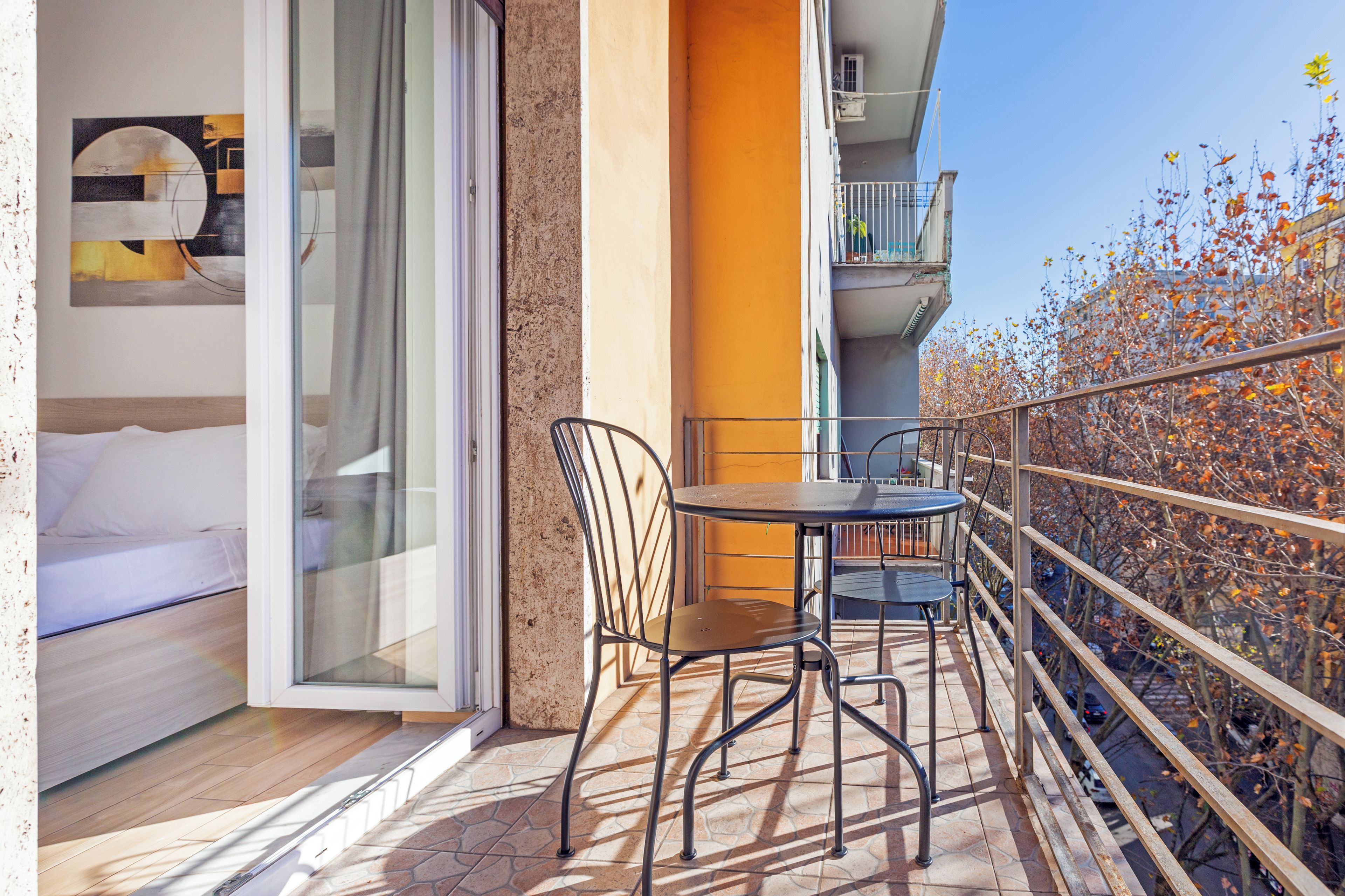 A balcony with a table and chairs, a bed, and a view of autumn trees.
