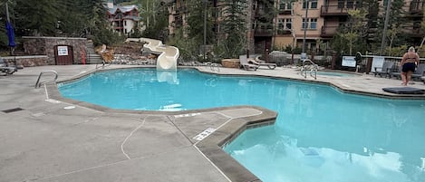 Una piscina con tobogán, rodeada por un paisaje montañoso y edificios de una cabaña.