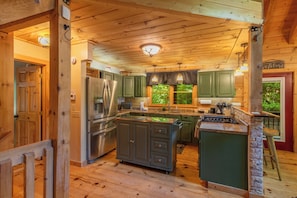 A cabin-style kitchen with wooden beams, a large island, stainless steel appliances, and a stone accent wall. - Nantahala Adventures- Hot tub- Firepit- Foosball Table (Bryson City)