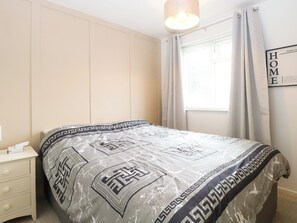 A bedroom with a bed, a nightstand, a window with curtains, and a wall-mounted light fixture. - Leaf Lodge (Great Yarmouth)