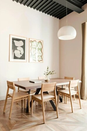 A dining area with a wooden table and chairs, framed artwork on the wall, and a hanging light fixture.
