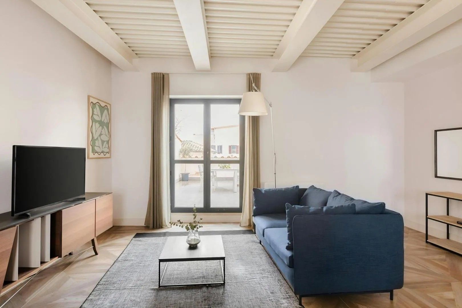 Una sala de estar moderna con un televisor de pantalla plana, un sofá azul oscuro, una mesa de centro y un amplio ventanal con cortinas.