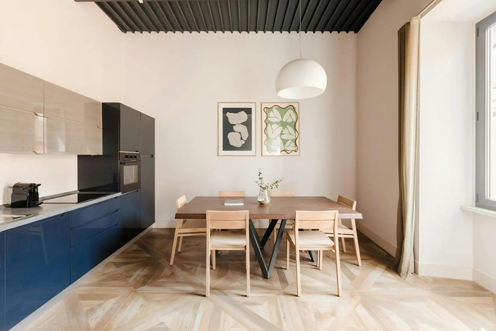 A modern kitchen with a dining area, featuring a wooden table and chairs, a hanging light fixture, and artwork on the wall.