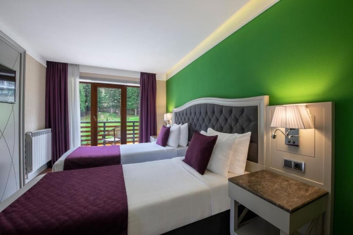 A hotel room with two beds, a nightstand, a lamp, and a view of the outdoors.