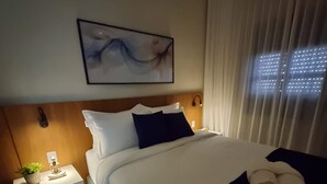 Ett hotellrum med en säng, ett nattduksbord, en tavla och ett fönster med gardiner.