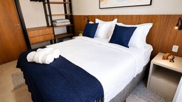 Quarto de hotel com uma cama, uma mesa de cabeceira, uma prateleira na parede e uma porta.
