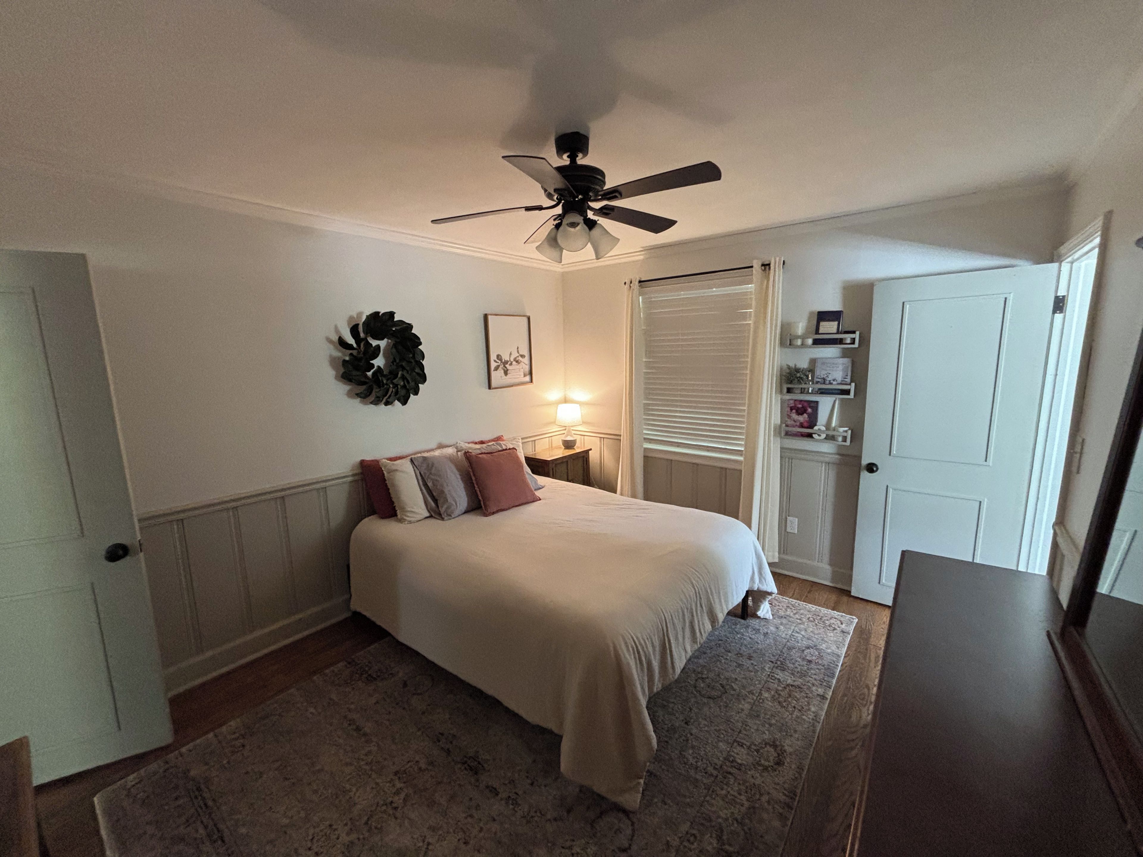 A bedroom with a bed, a ceiling fan, a wall-mounted wreath, a bedside table with a lamp, and a door with a mirror.
