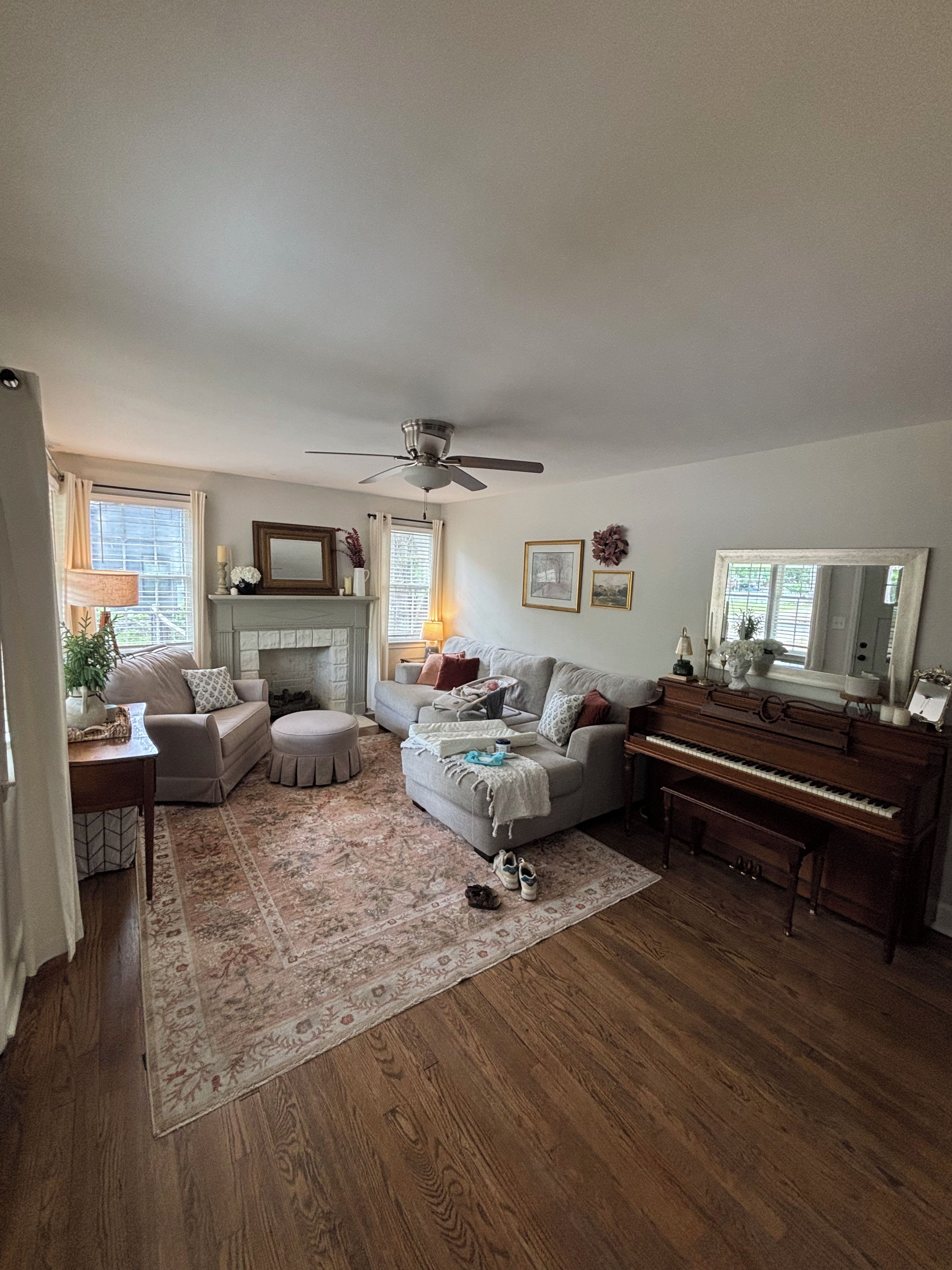 A living room with a fireplace, a ceiling fan, a piano, and a large window with curtains.