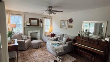 A living room with a fireplace, a ceiling fan, a piano, and a large window with curtains.