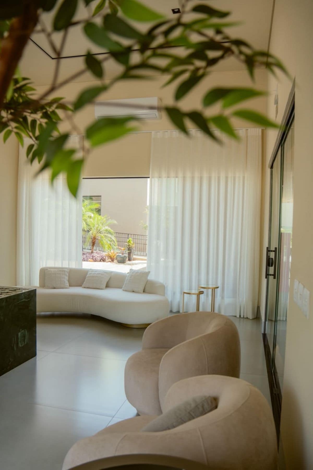 Uma sala de estar moderna com um sofá curvo, uma mesa de centro de vidro e vista para uma varanda com plantas.