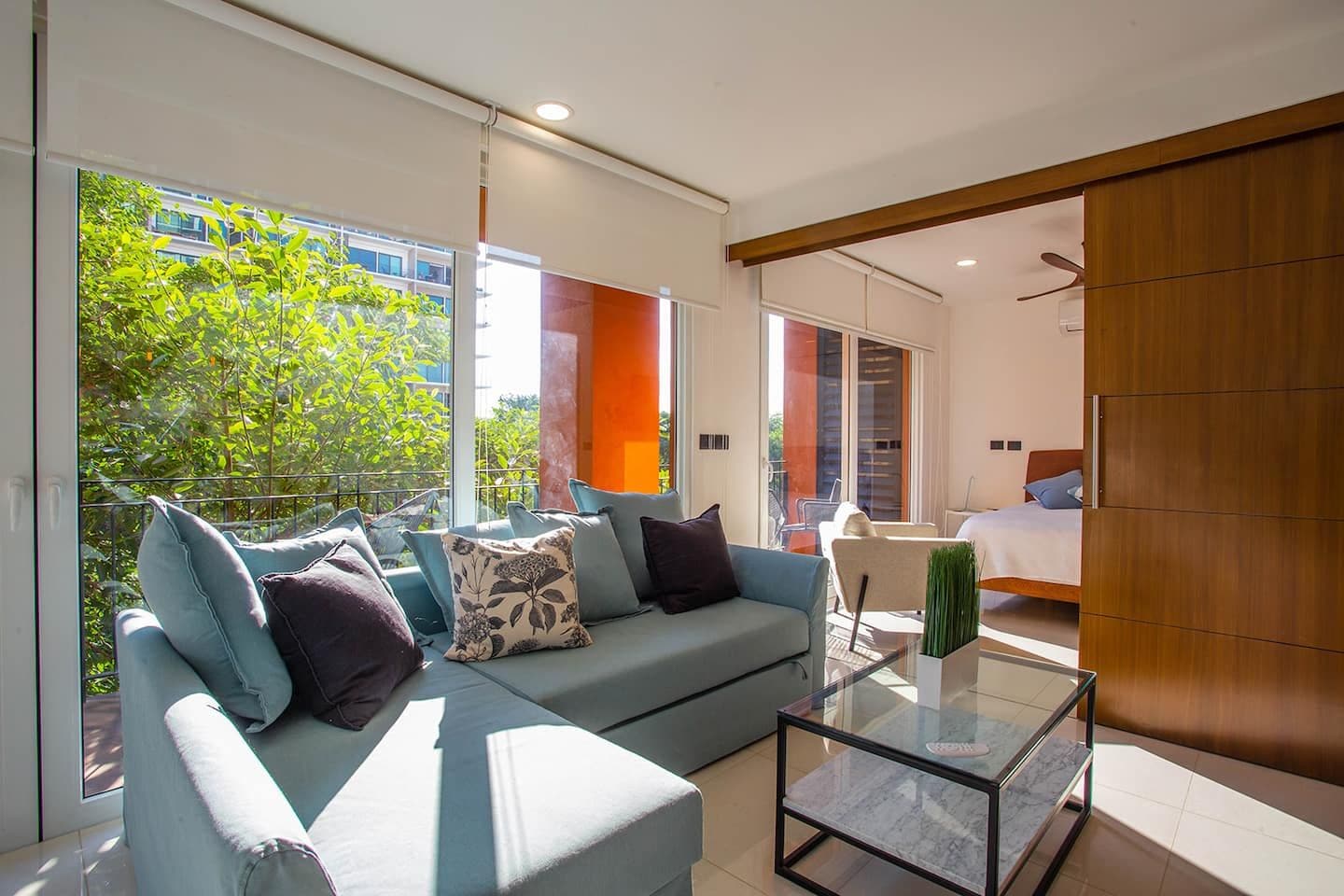 A modern living room with a sofa, a glass coffee table, and a view of a balcony with greenery.
