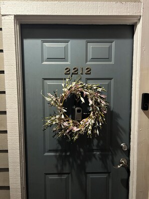 Une porte sombre, portant le numéro 2212, une sonnette métallique et une couronne de fleurs.