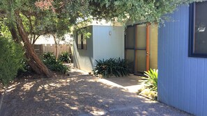A small, single-story building with a covered patio area, surrounded by various plants and a tree. - Beach Blue - Point Lonsdale (POINT LONSDALE)