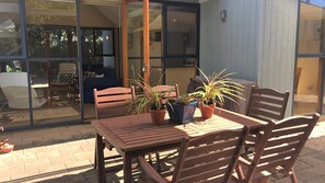 An outdoor patio with a wooden table and chairs, potted plants, and a glass door leading to an interior space. - Beach Blue - Point Lonsdale (POINT LONSDALE)