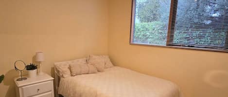 A bedroom with a single bed, a bedside table with a lamp, and a window with blinds.