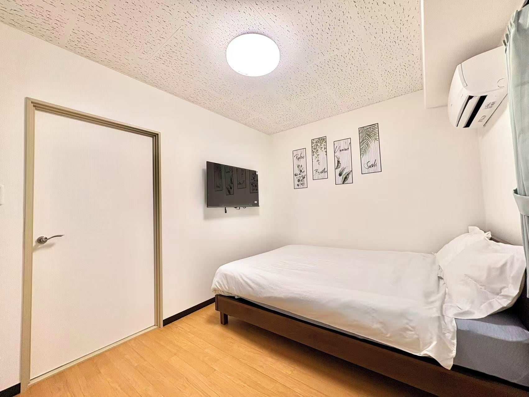 A bedroom with a bed, a door, a wall-mounted TV, and a ceiling light.