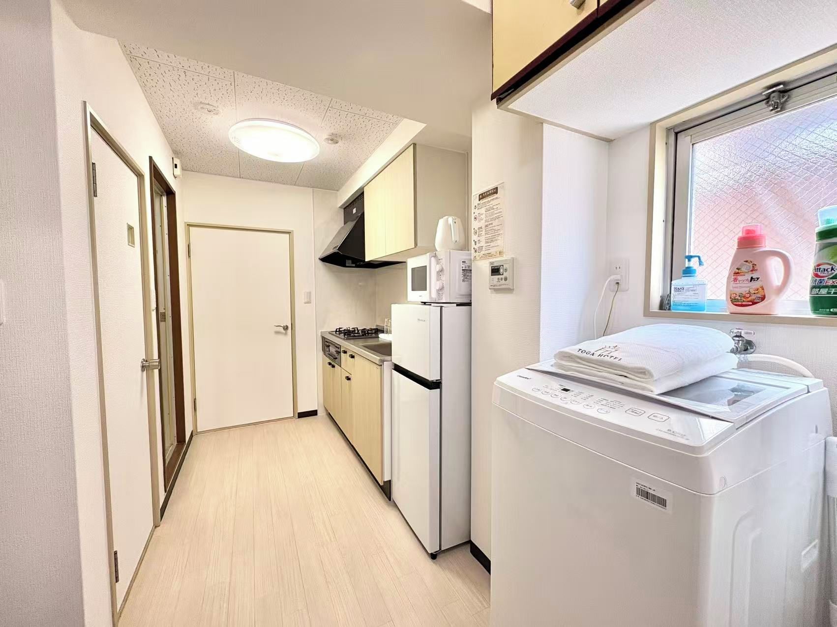 A compact hotel room with a kitchenette, a washing machine, and a window with cleaning supplies.