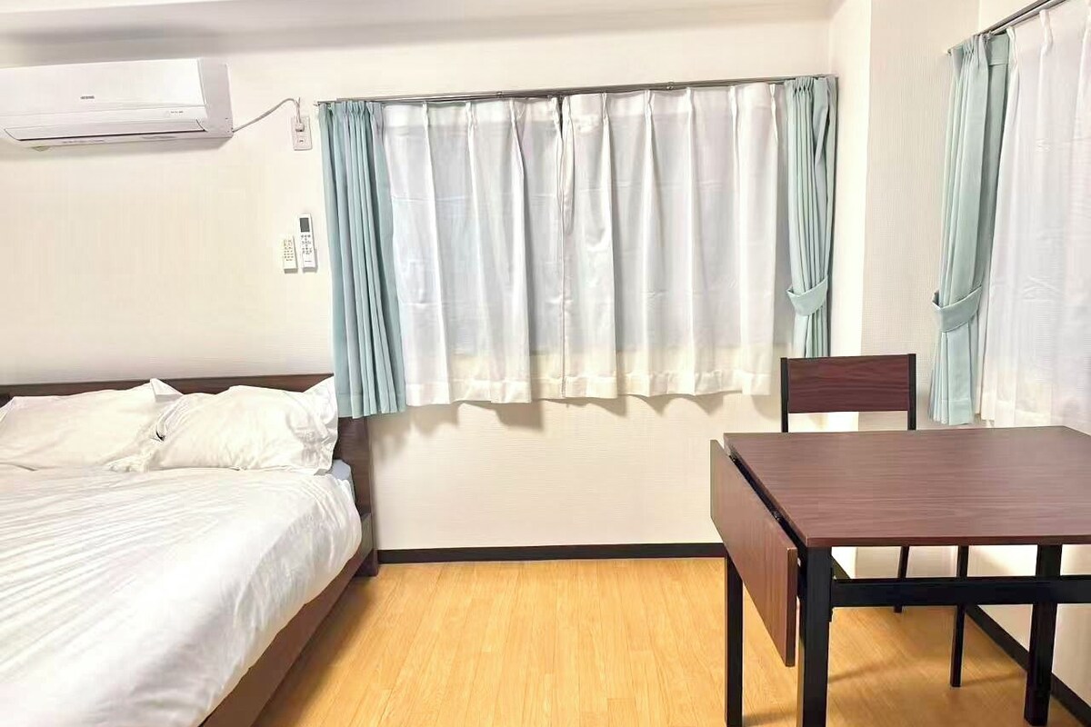 A room with a bed, a table, and a chair. There is a window with curtains.