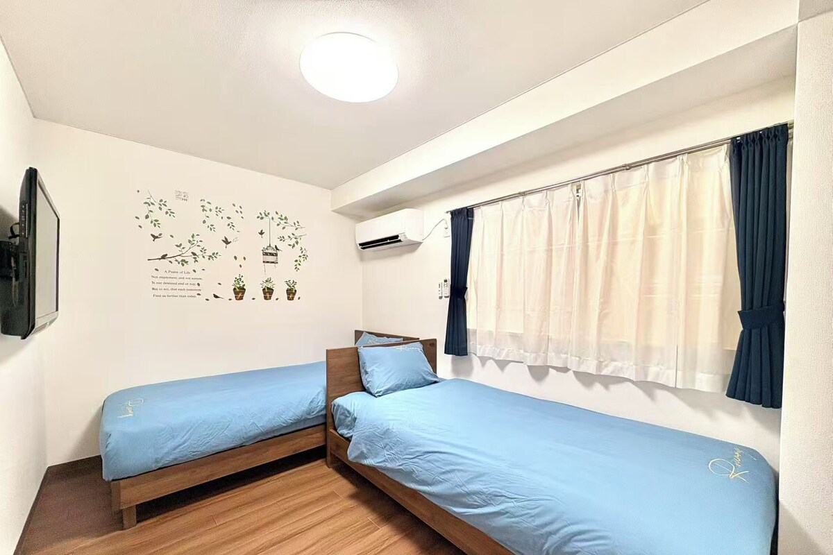 A room with two single beds, a ceiling light, an air conditioning unit, and a window with curtains.