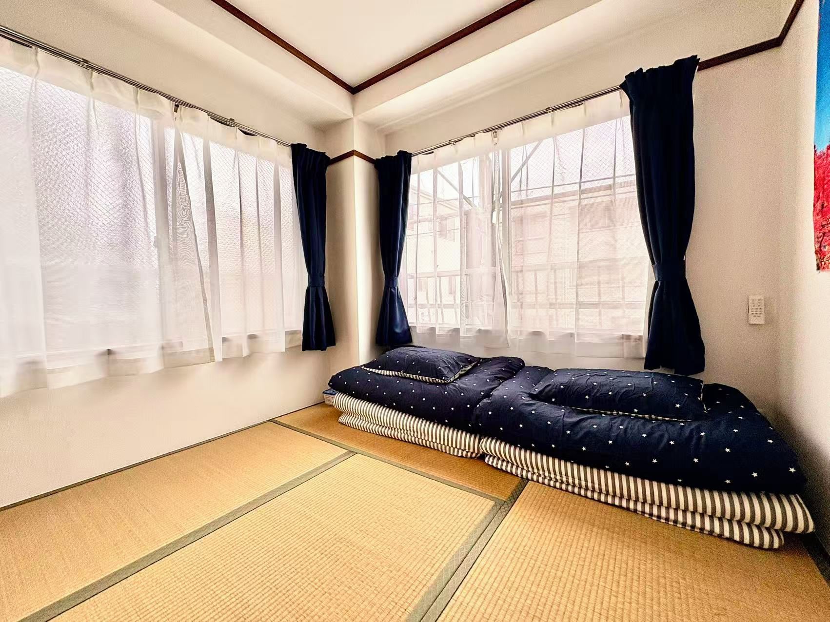 A room with tatami flooring, a bed with a dark patterned bedspread, and sheer curtains on the windows.