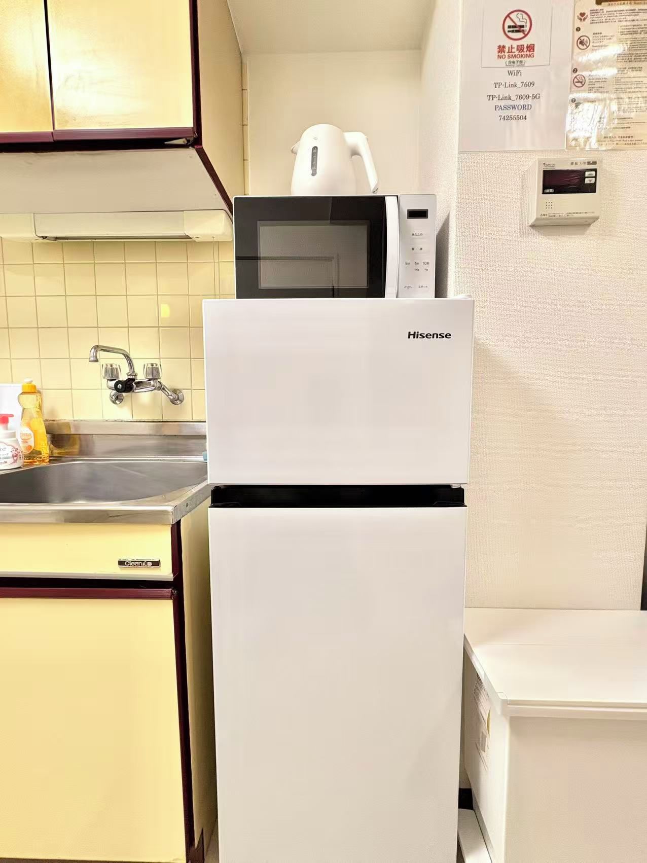 Una cocina con un refrigerador blanco, un microondas y una tetera. Hay un grifo y una botella de jugo de naranja en la encimera.