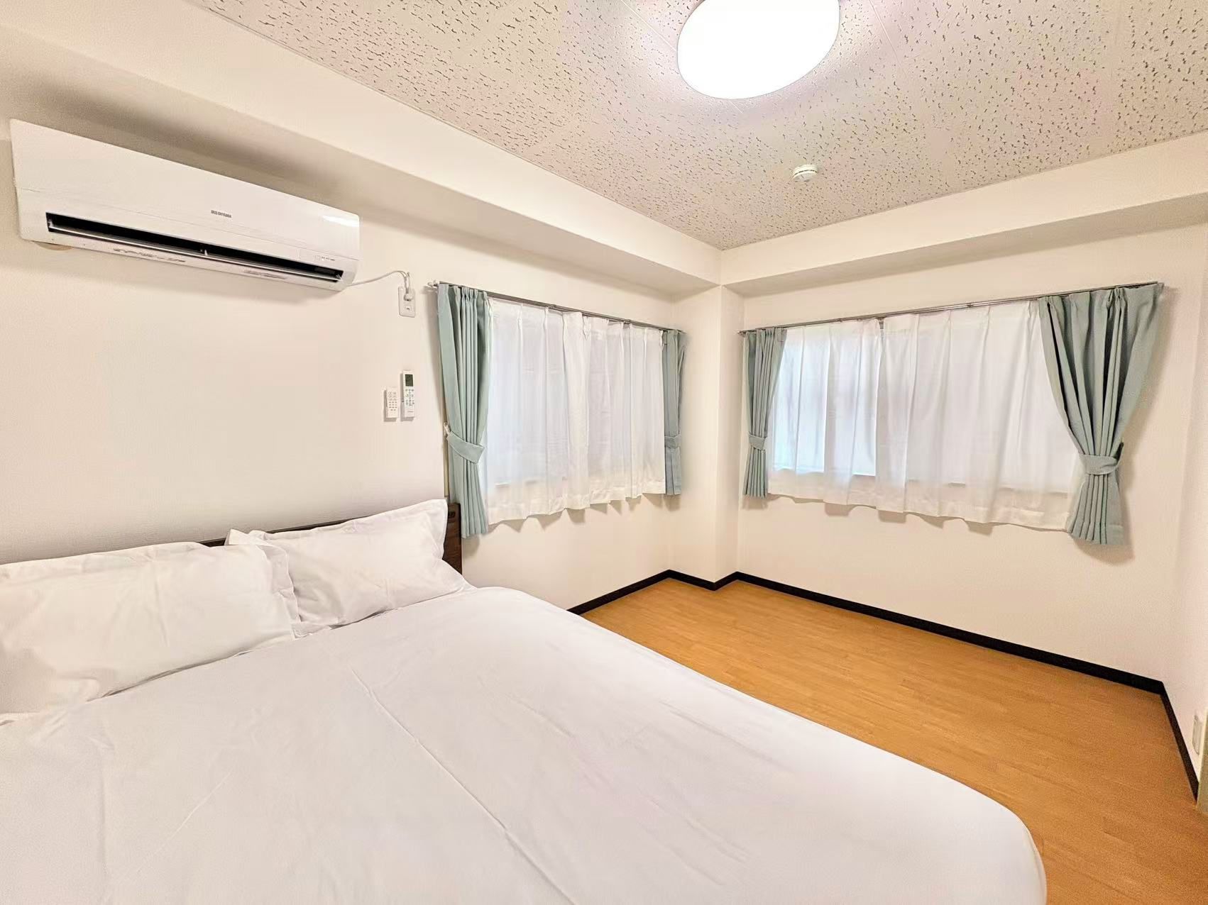 A hotel room with a bed, an air conditioning unit, two windows with curtains, and a wooden floor.