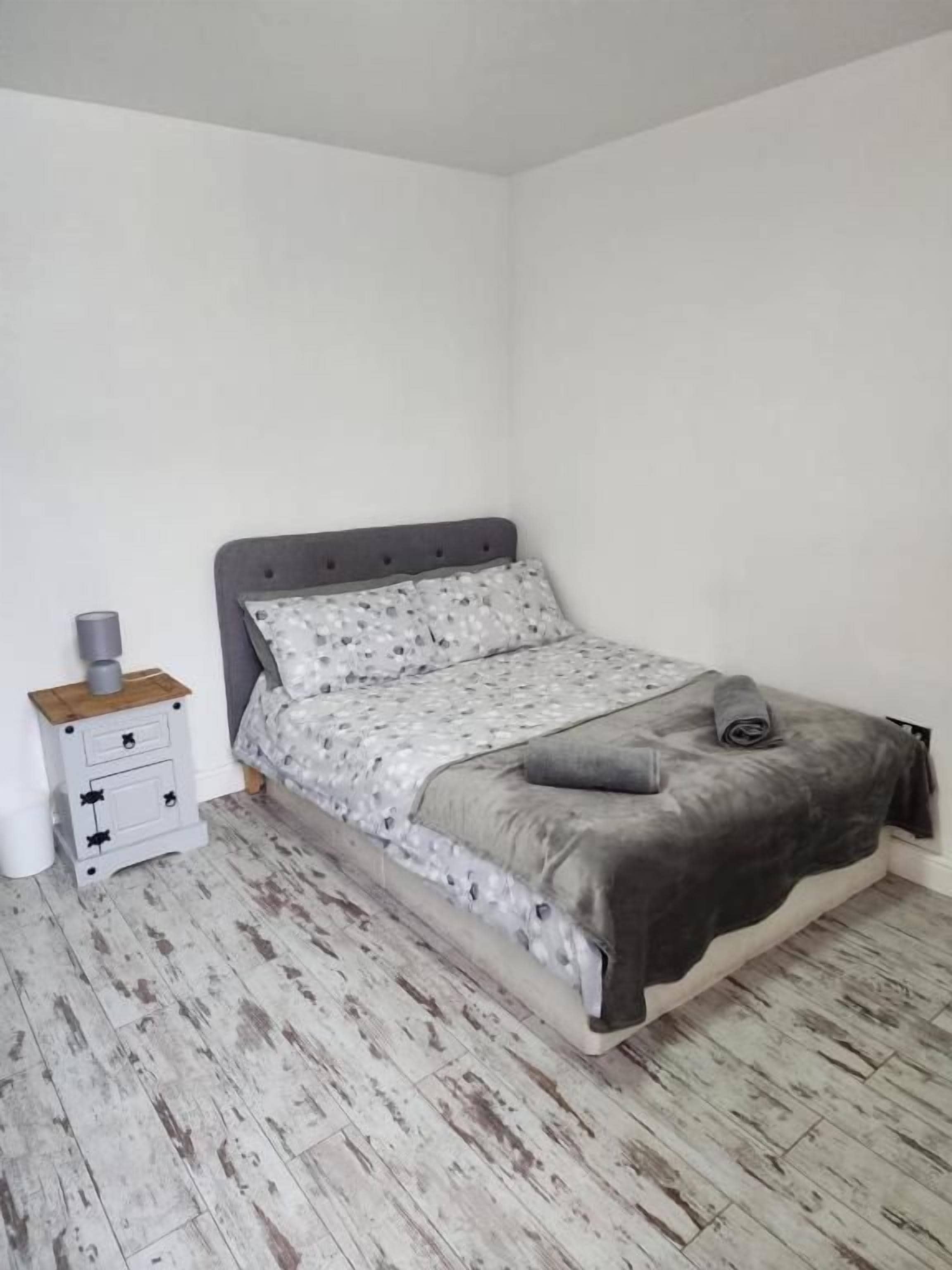 A single bed with a grey headboard, a patterned bedspread, and a grey blanket. There is a small bedside table with drawers and a lamp. The floor is wooden with a distressed finish.