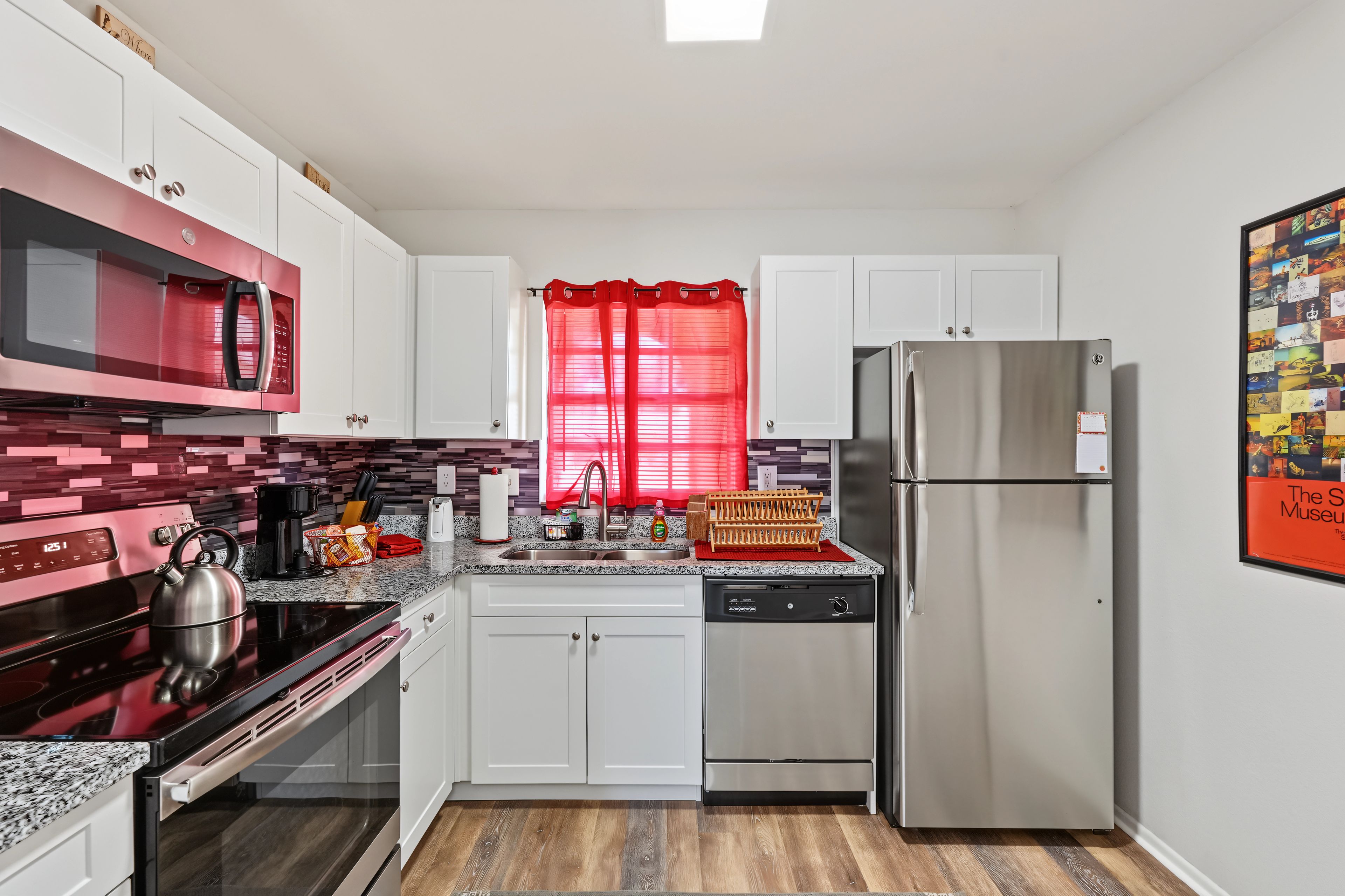 A modern kitchen with white cabinets, stainless steel appliances, and a red window curtain.
