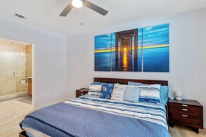 Ein Schlafzimmer mit Bett, Nachttischen, einem Deckenventilator und einem großen Gemälde eines Piers bei Sonnenuntergang.