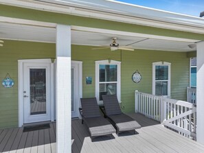 A covered porch with two lounge chairs, a white door, and a ceiling fan. - Mer Sea, 4 bedroom 2.5 bath home sleeps 13. A short stroll to the beach. (Crystal Beach)