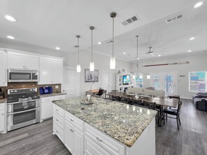 Une cuisine moderne avec des meubles blancs, un îlot en granit, des appareils en acier inoxydable, un éclairage suspendu et un coin repas avec une grande table et des chaises.
