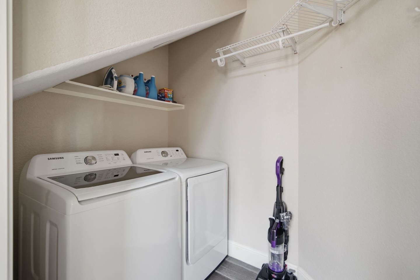 A laundry room with two white washers and dryers, a shelf with cleaning supplies, and a vacuum cleaner.