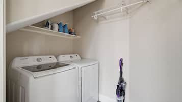 A laundry room with two white washers and dryers, a shelf with cleaning supplies, and a vacuum cleaner.